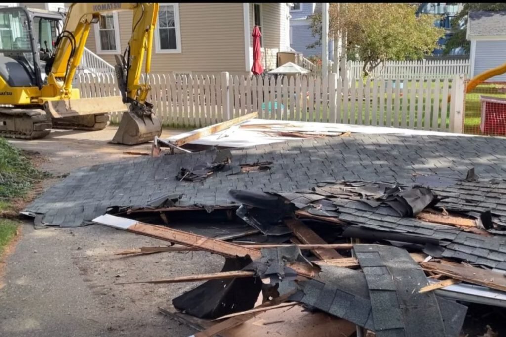 Excavator tearing down a small structure with the collapsed roof and shingle debris ready to be hauled out