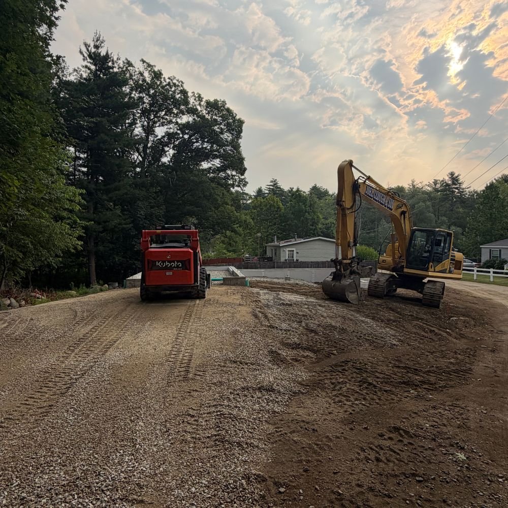 Skid steer and excavator grading a gravel driveway and parking area at dusk