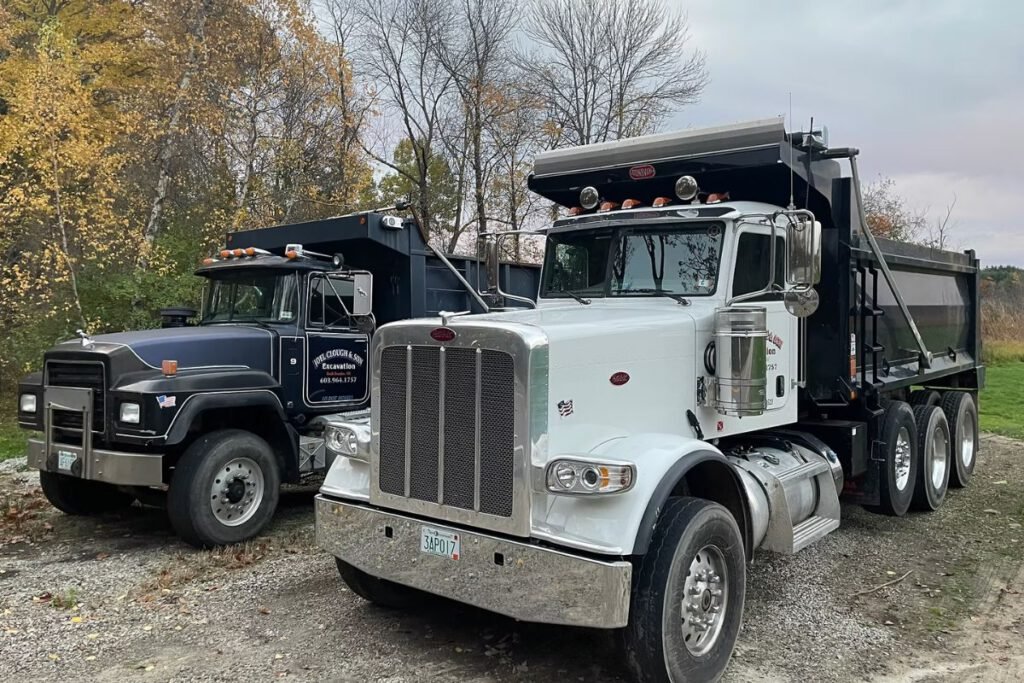 Two Joel Clough and Son dump trucks parked on a gravel yard, used for hauling loam, gravel and fill