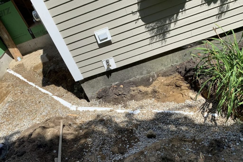 White drain line bedded in crushed stone running along a house foundation before backfill