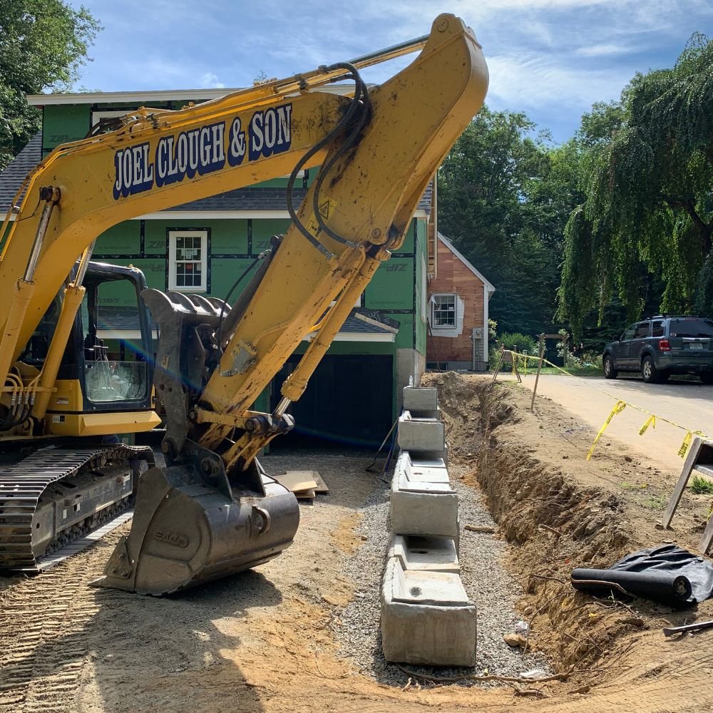 Joel Clough and Son branded excavator working beside a house under construction with concrete blocks lining a new trench